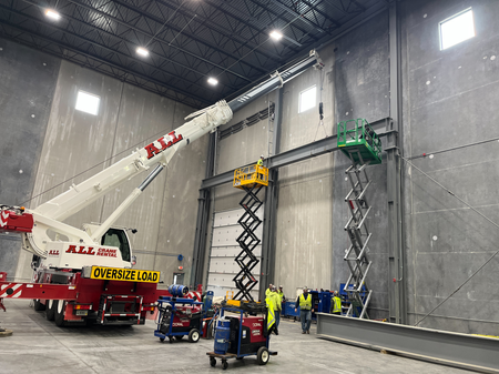 Crane lifting a steel beam in a large industrial building. Workers on lifts are near the beam and welding equipment is present.