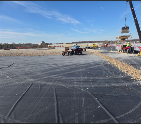 Workers spreading gravel on a black surface with ATVs, crane, and bucket on a sunny day.