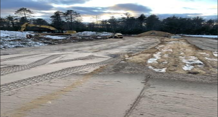 Muddy construction road with tire tracks, snow piles, and trees under a cloudy sky