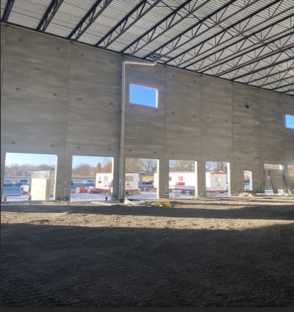 Interior view of a concrete wall under construction with window openings, metal roof structure and gravel floor.