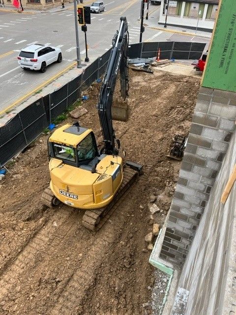 Yellow John Deere excavator on construction site. Road, car, and buildings in background.