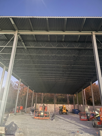 Construction site with a partially built metal roof supported by steel beams. Various construction vehicles are present.
