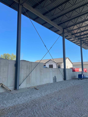 Steel support beams with diagonal bracing along a concrete wall, under a metal roof, against a blue sky.