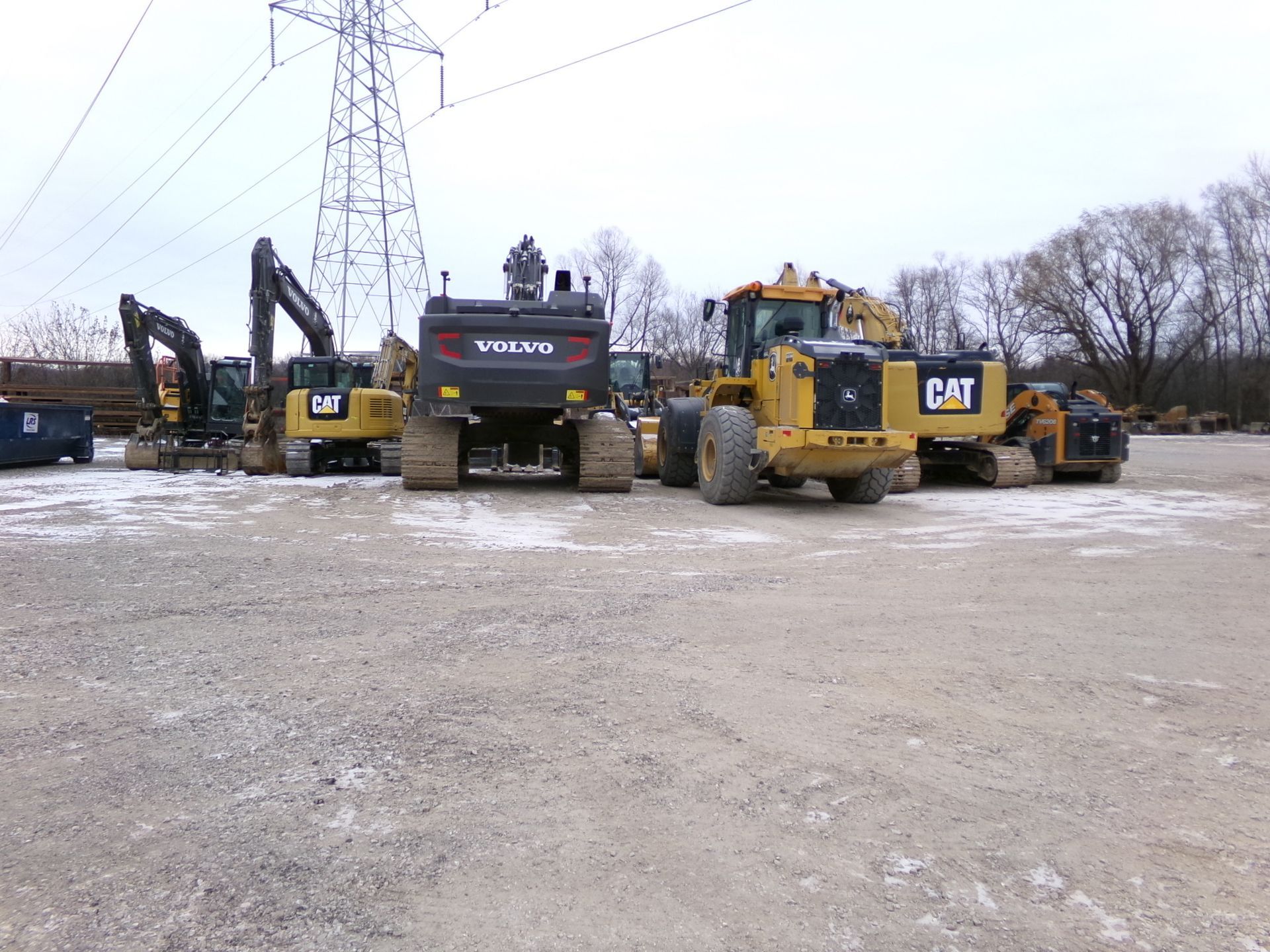 Construction equipment, including CAT and Volvo, parked on gravel in front of power lines.