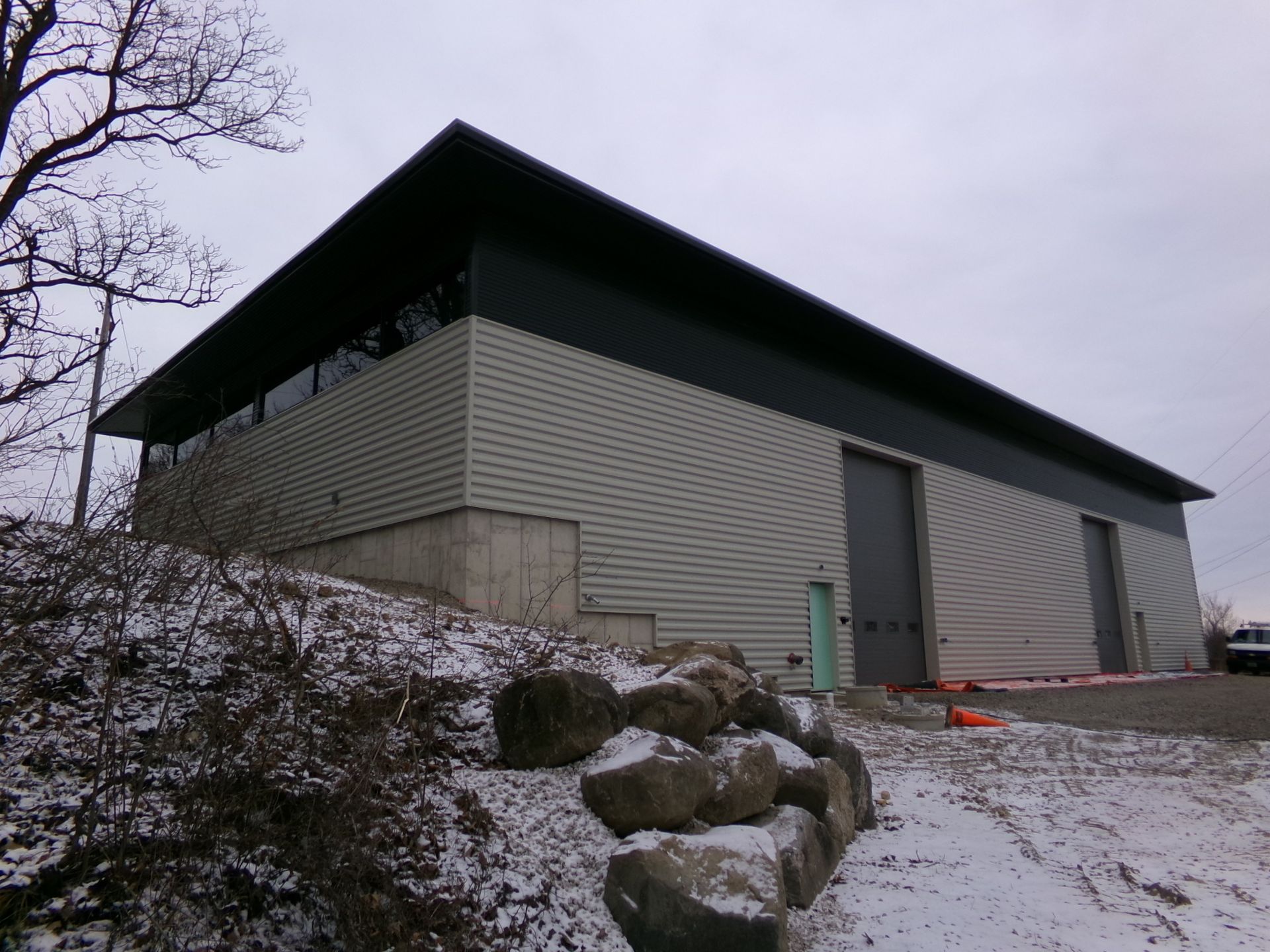 Gray industrial building with dark roof, two garage doors, snow, and large rocks in front.