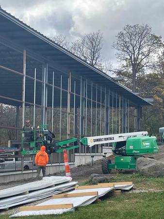 Construction of a building with glass panels. Workers on a lift, wearing orange vests. Sheets of material on the ground.