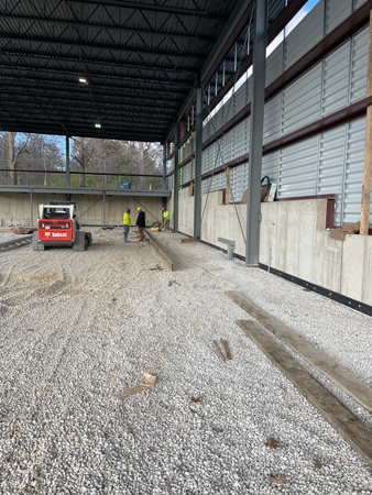 Construction site: Gravel ground, Bobcat, workers, and a partially built metal building with concrete walls.