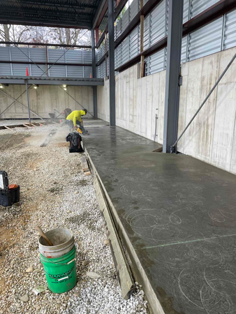 Construction site with worker smoothing wet concrete floor. Gray walls, gravel, and metal beams are visible.