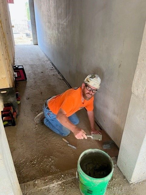 Construction worker kneeling, applying material to a floor; orange shirt, bandana, bucket of material.