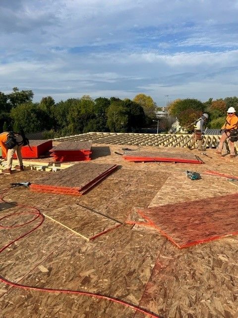 Workers on a rooftop install wood sheeting. Construction site with blue sky, trees, and scattered materials.
