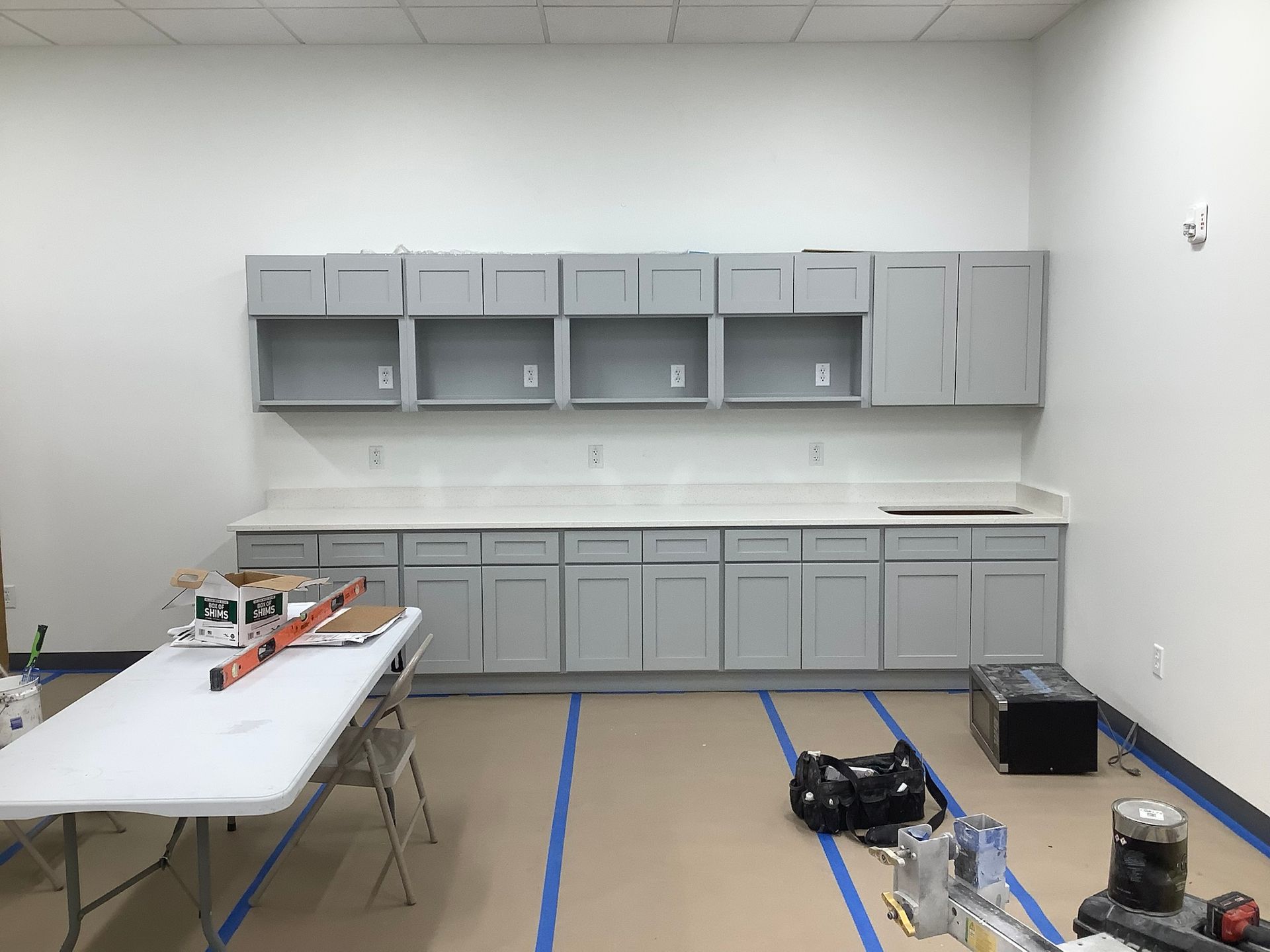 Gray cabinets along white wall with white countertop. Construction tools and table in foreground.