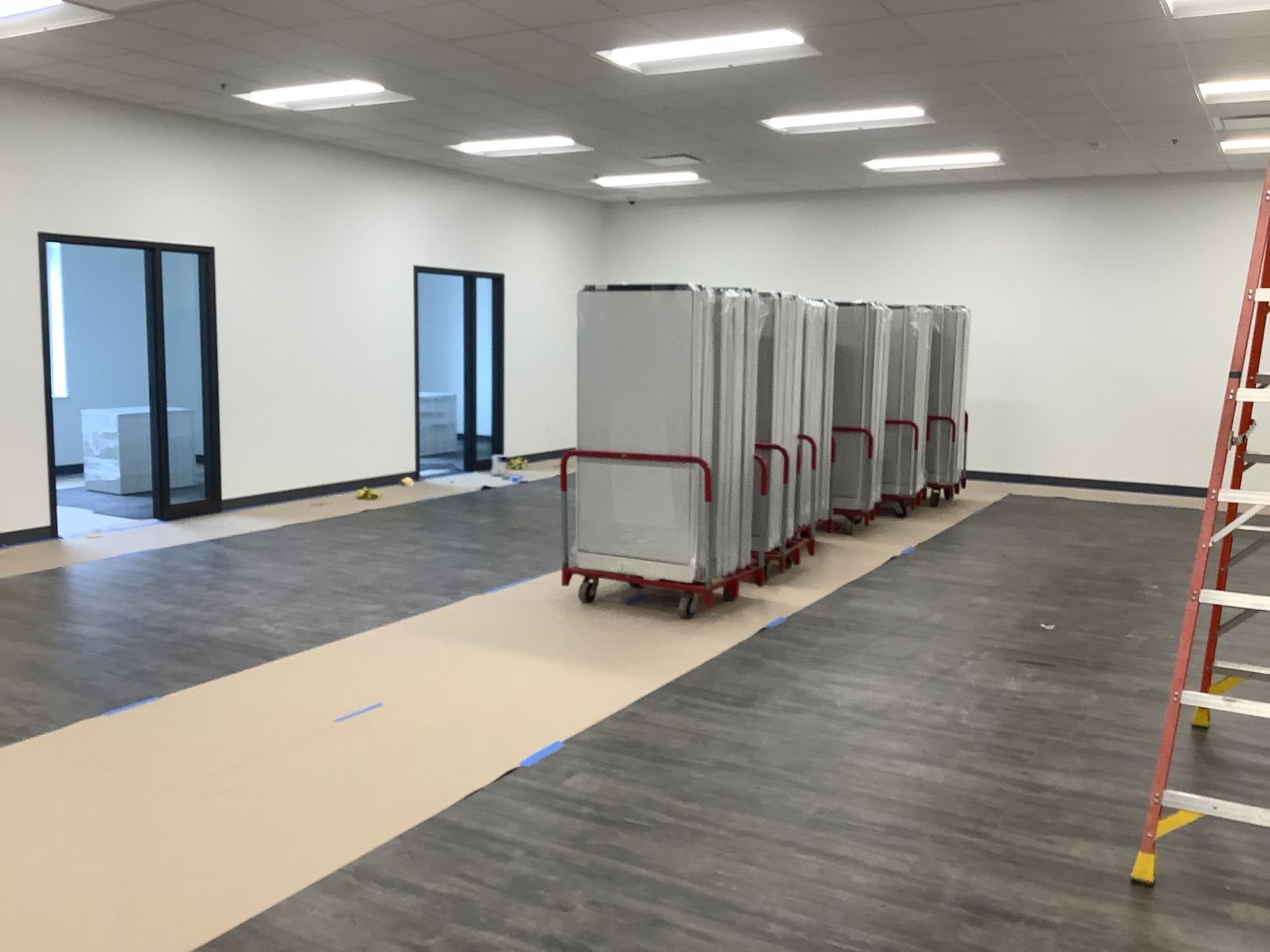 Empty room with gray flooring, white walls, and carts holding panels. A ladder is present.