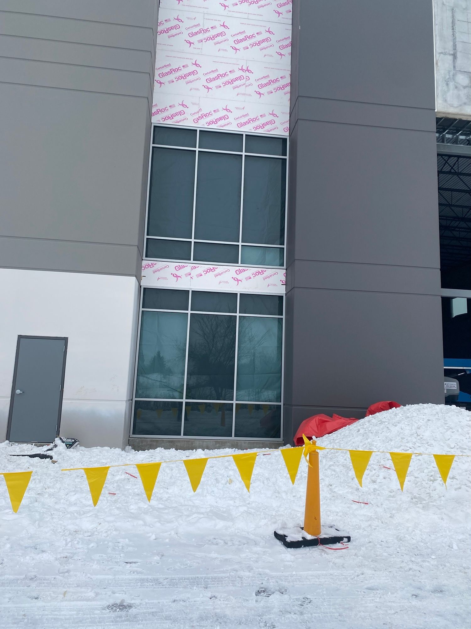 Building exterior with windows under construction, snowy ground, yellow caution flags.