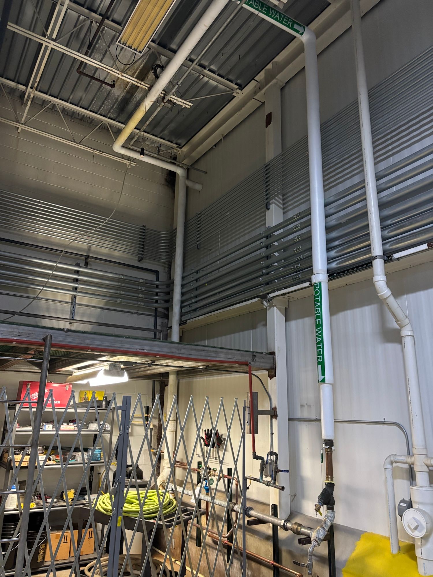 White pipes run vertically and horizontally across a ceiling, near a wall and a cluttered work area.