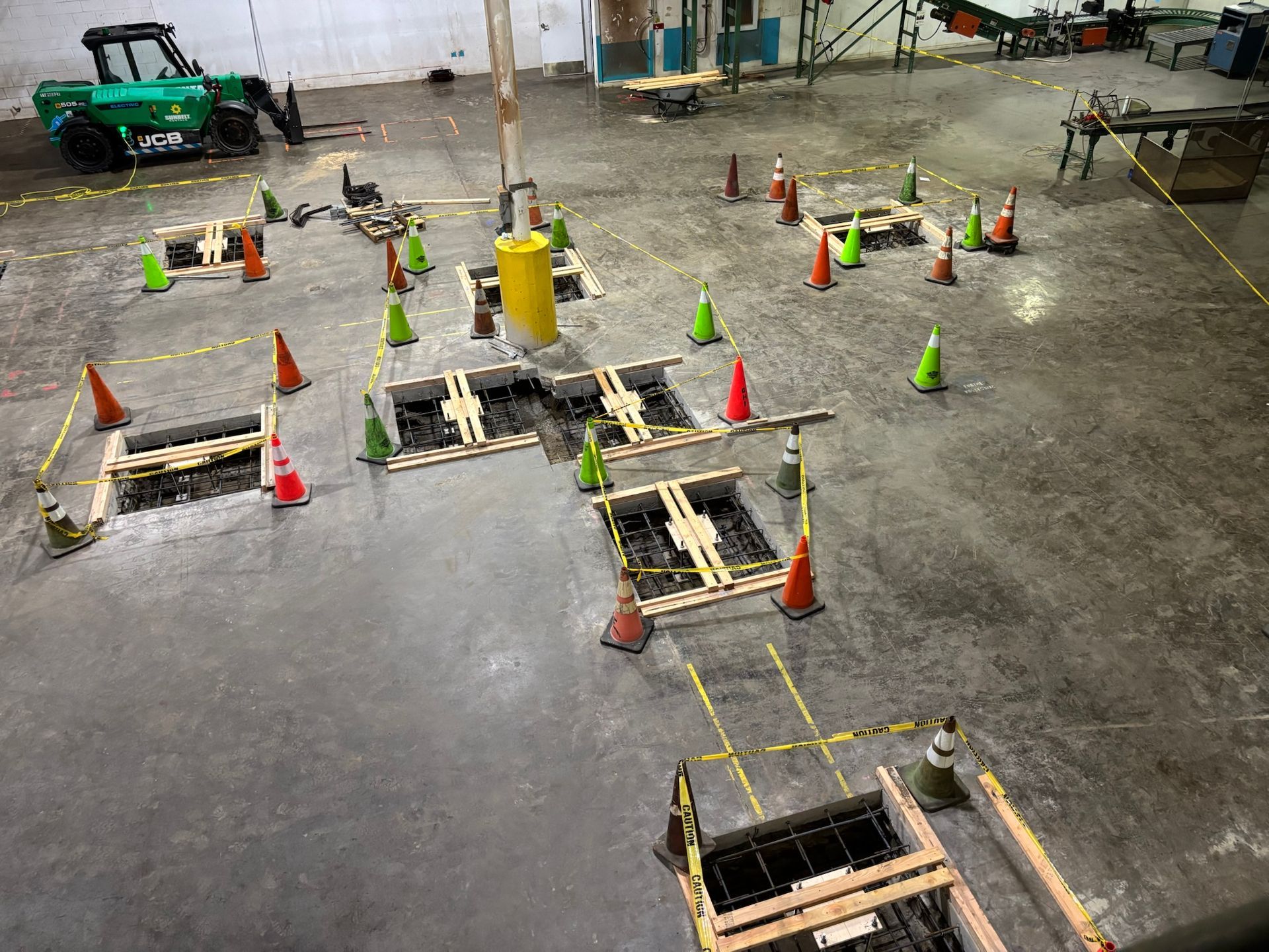 Indoor construction site with marked floor openings, orange cones, and a green machine in the background