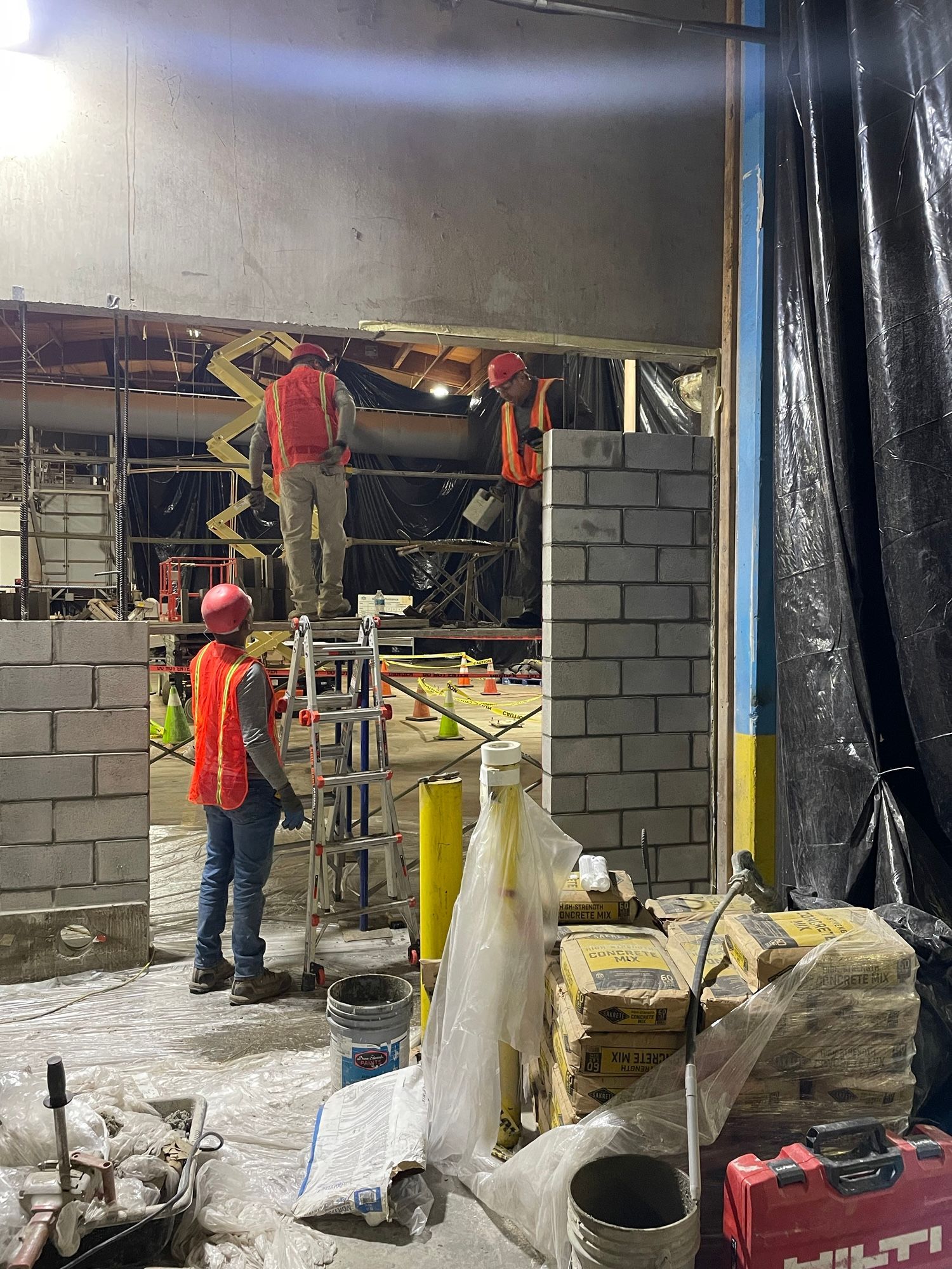 Workers building a cinder block wall inside a construction site, with scaffolding and tools around them.