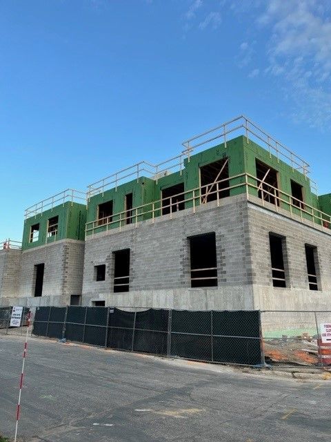 Construction site: two-story building in progress, grey cinder block walls, green roof supports, safety barriers.