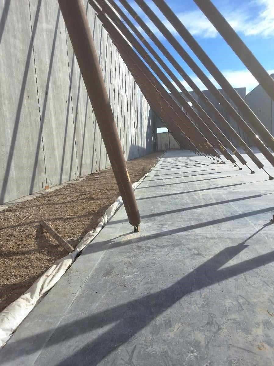 Concrete walkway with large wooden beams angled against a textured concrete wall, lit by sunlight.