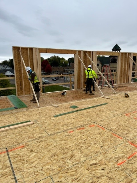 Construction workers framing a building's exterior wall on a rooftop, bracing the wooden structure with temporary supports.