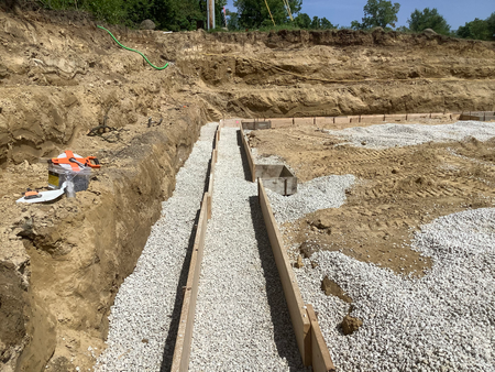 Foundation construction site with gravel-filled trenches lined with wooden forms, on a sunny day.