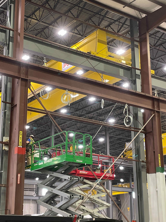 Worker on a green scissor lift, near brown steel beams. Yellow industrial crane visible in large warehouse.