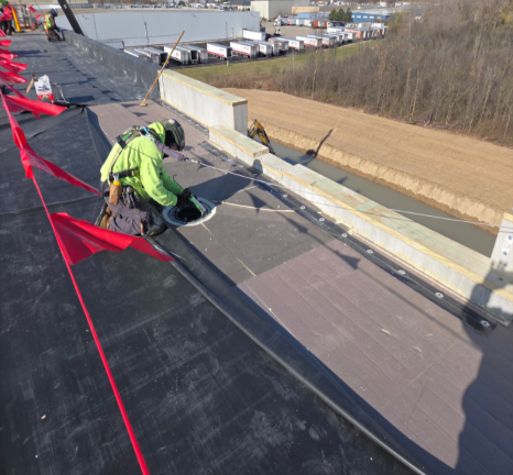Roofer in safety gear near a drain on a black roof, near a concrete barrier. Red flags attached.