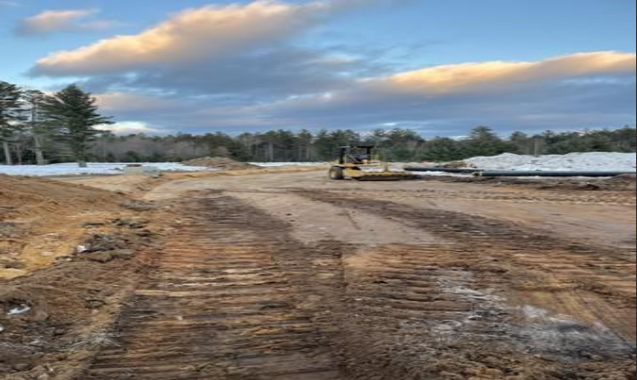 Dirt road construction site with heavy machinery under a cloudy sky, snow along the sides.