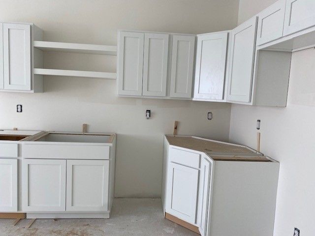 White kitchen cabinets installed in a room with unfinished walls and flooring, awaiting countertops.