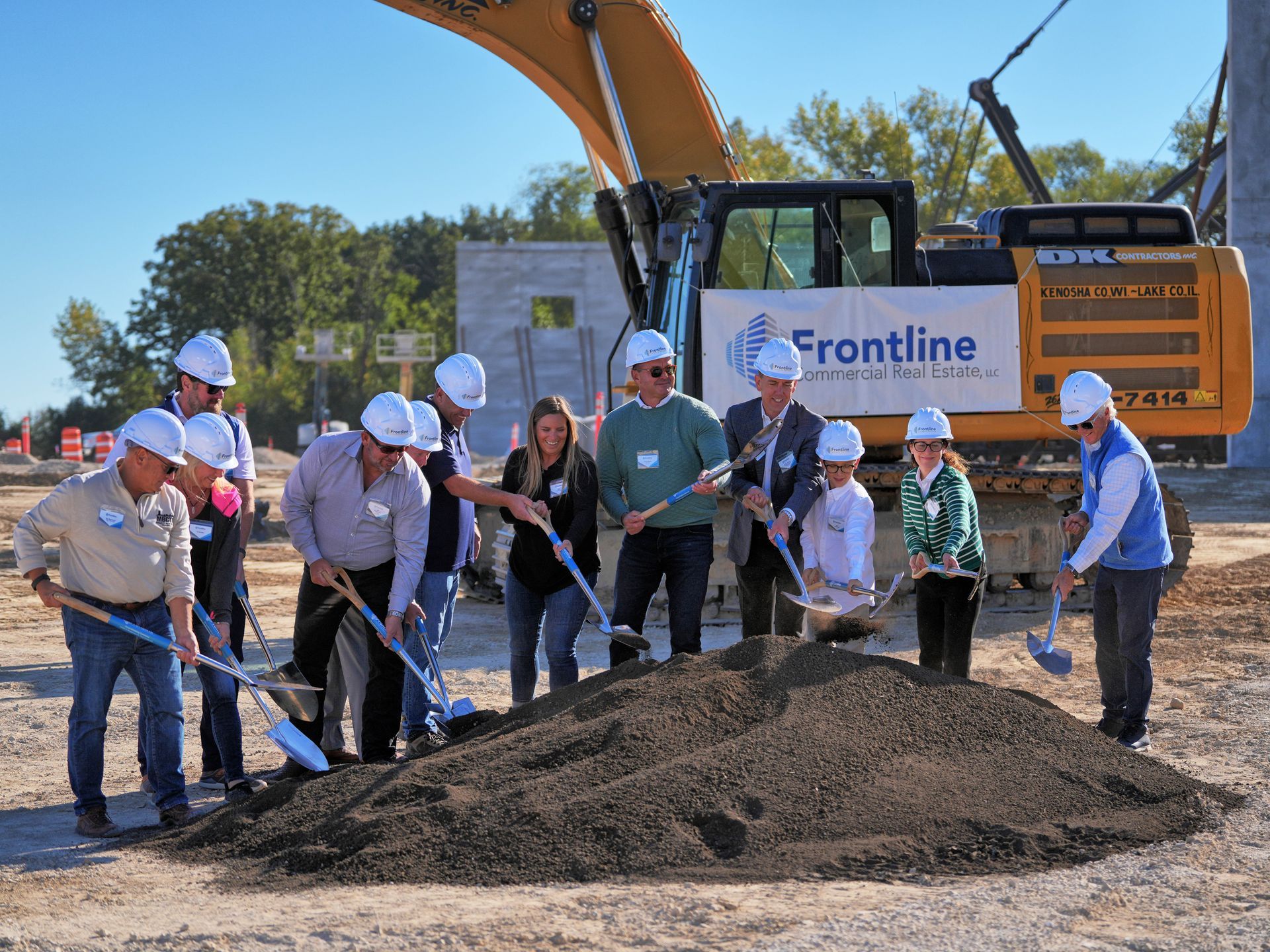 People in hard hats breaking ground with shovels near an excavator. 