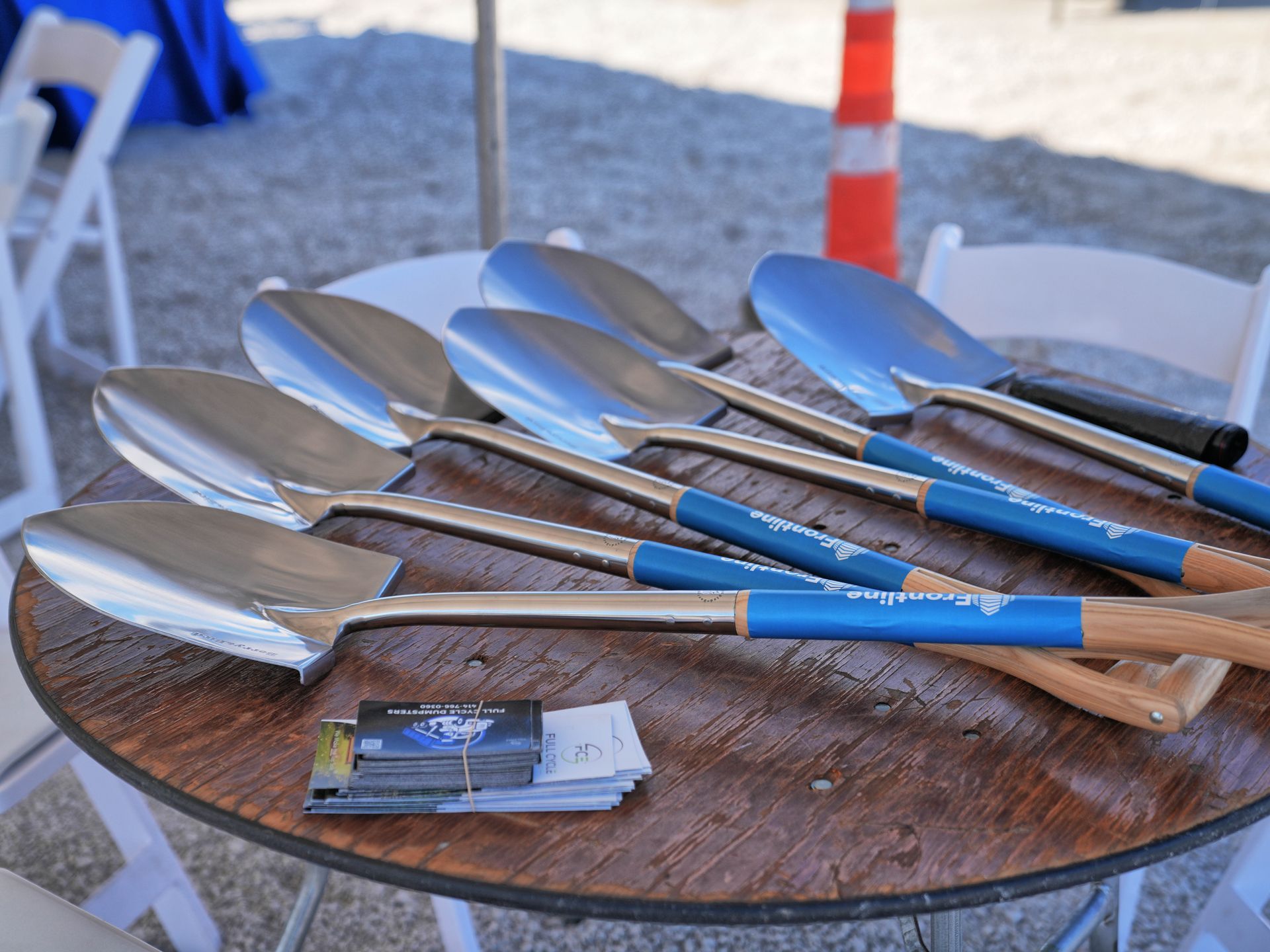 Six shiny shovels with blue handles on a wooden table, ready for a groundbreaking ceremony.