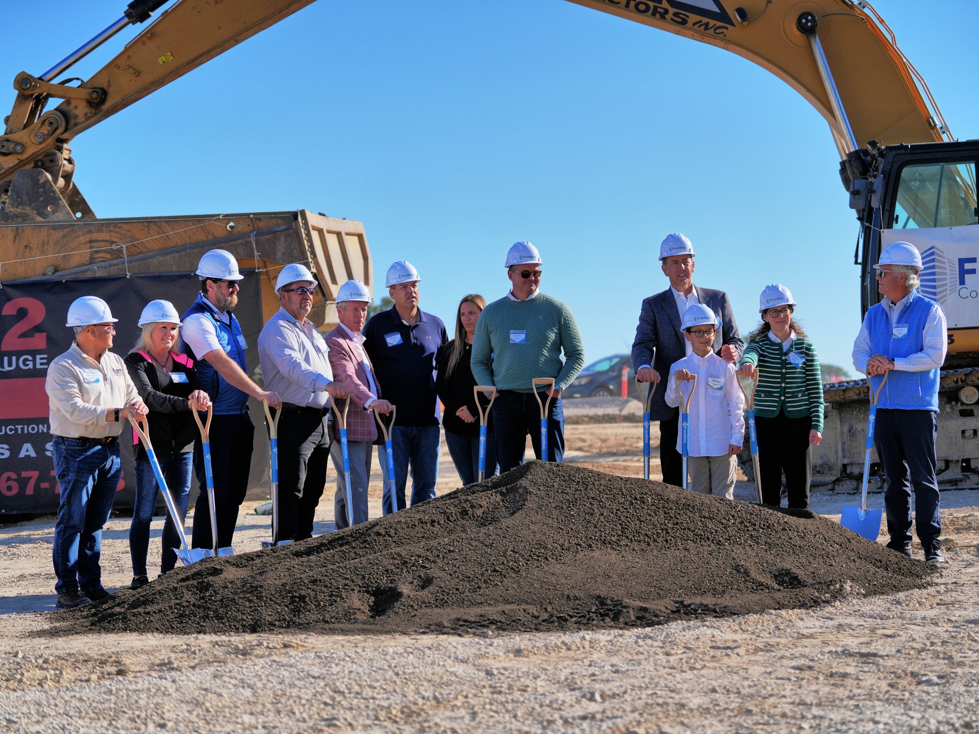 People with shovels at a groundbreaking ceremony, standing in front of a pile of dirt and an excavator.