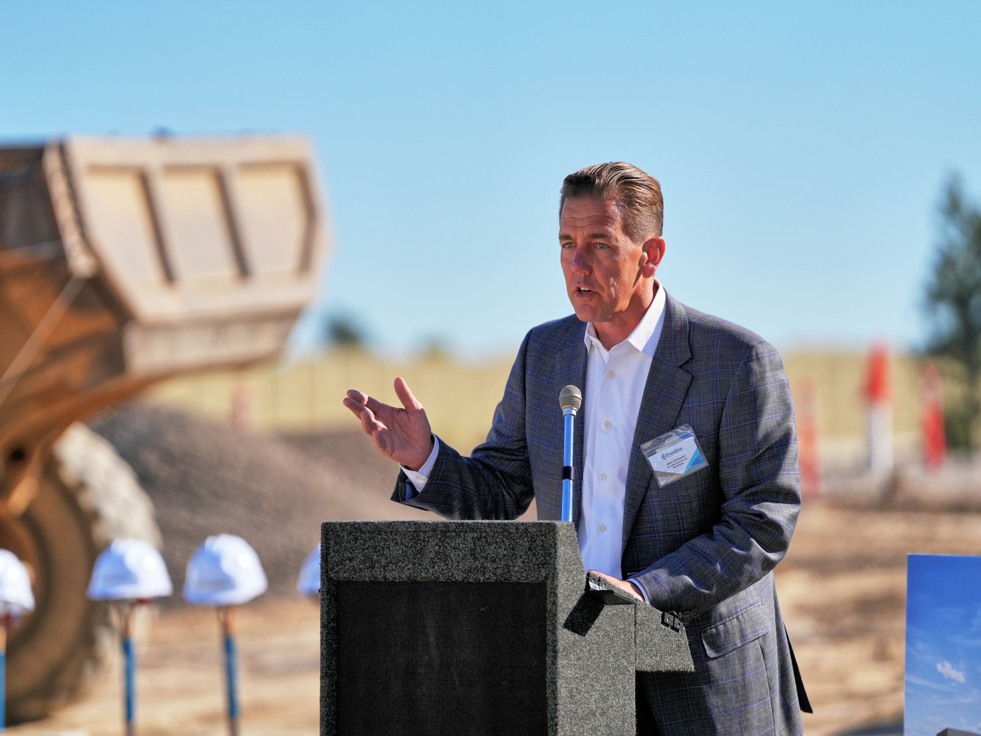 Man speaking at a podium with construction equipment in the background. Sunny outdoor setting.