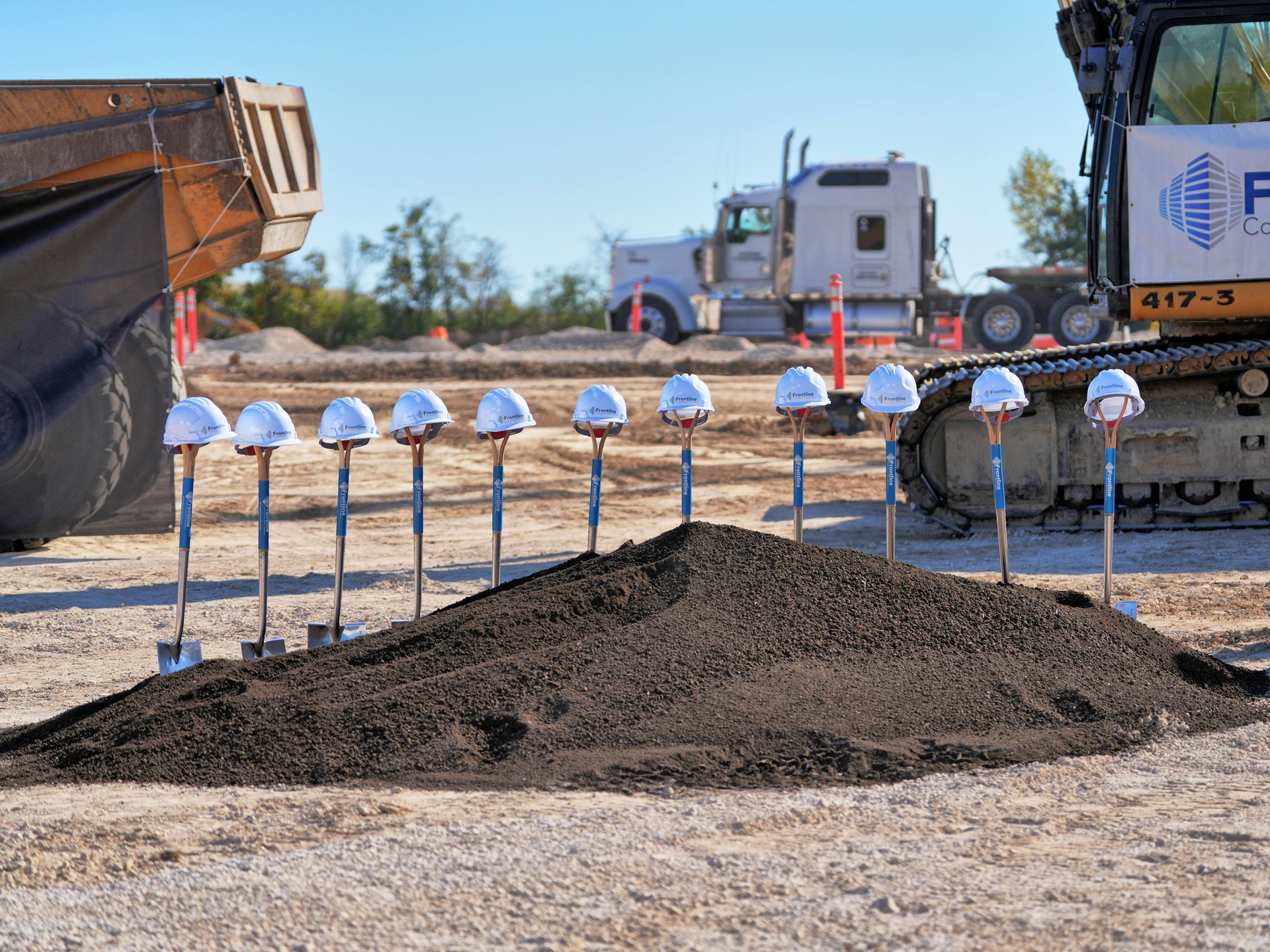 Groundbreaking ceremony with shovels and white hard hats. Construction equipment and truck in the background.