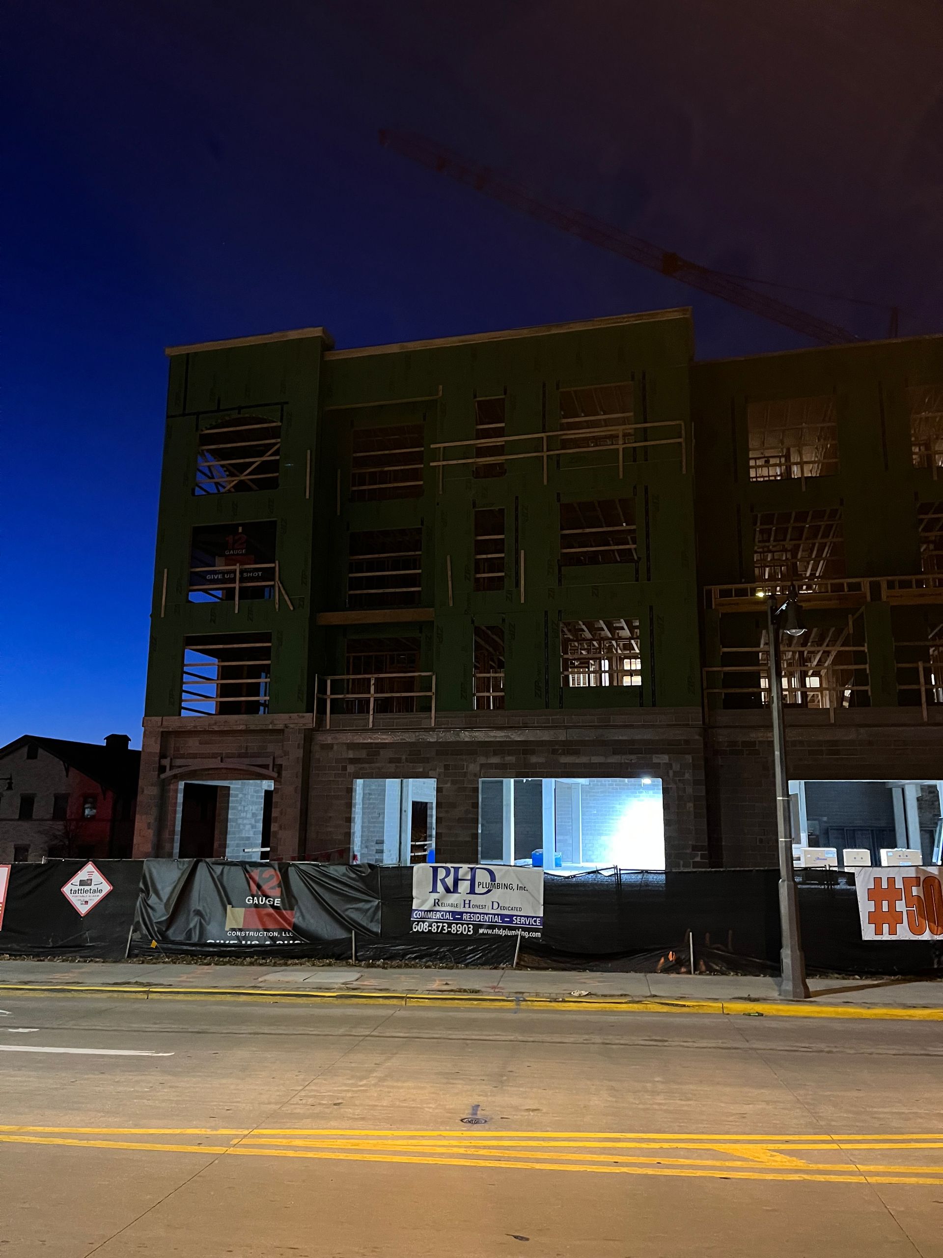 Building under construction at night, covered in green sheeting, with bright interior lights and black fencing.