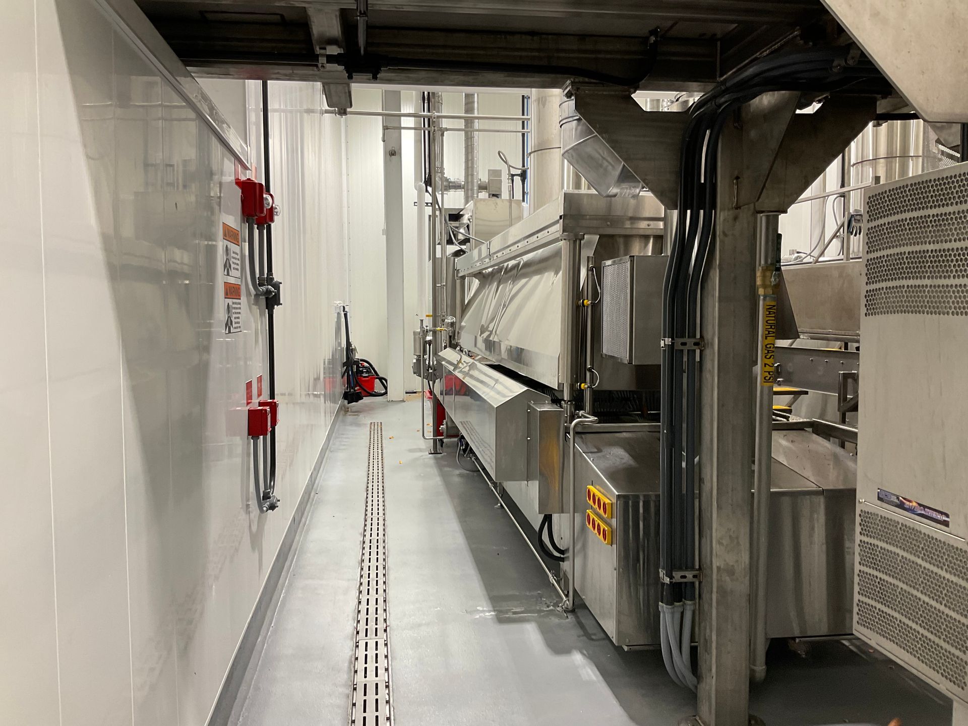 A narrow industrial hallway with machinery and electrical panels. White walls, grey floor.