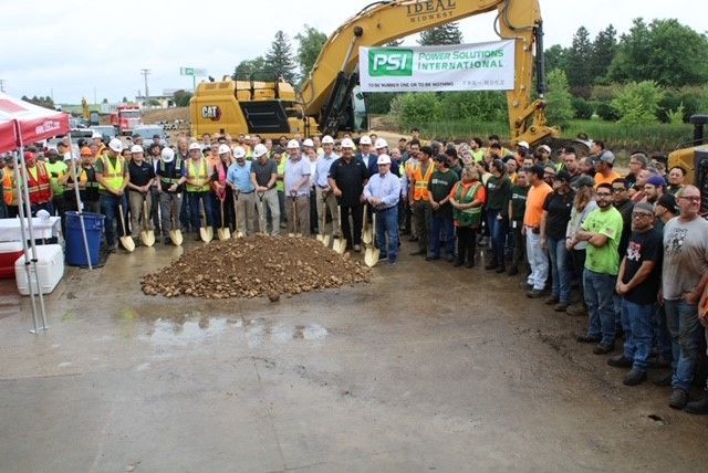 Groundbreaking ceremony with a large group holding shovels, yellow excavator in background.
