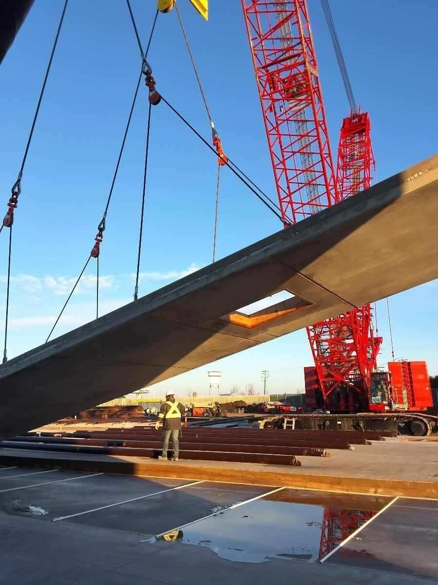 A large crane lifting a concrete beam. A worker in a vest watches. Blue sky.