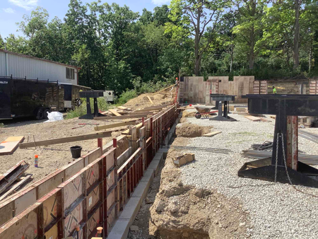 Construction site with concrete forms, gravel, and a partially built structure, trees in the background.