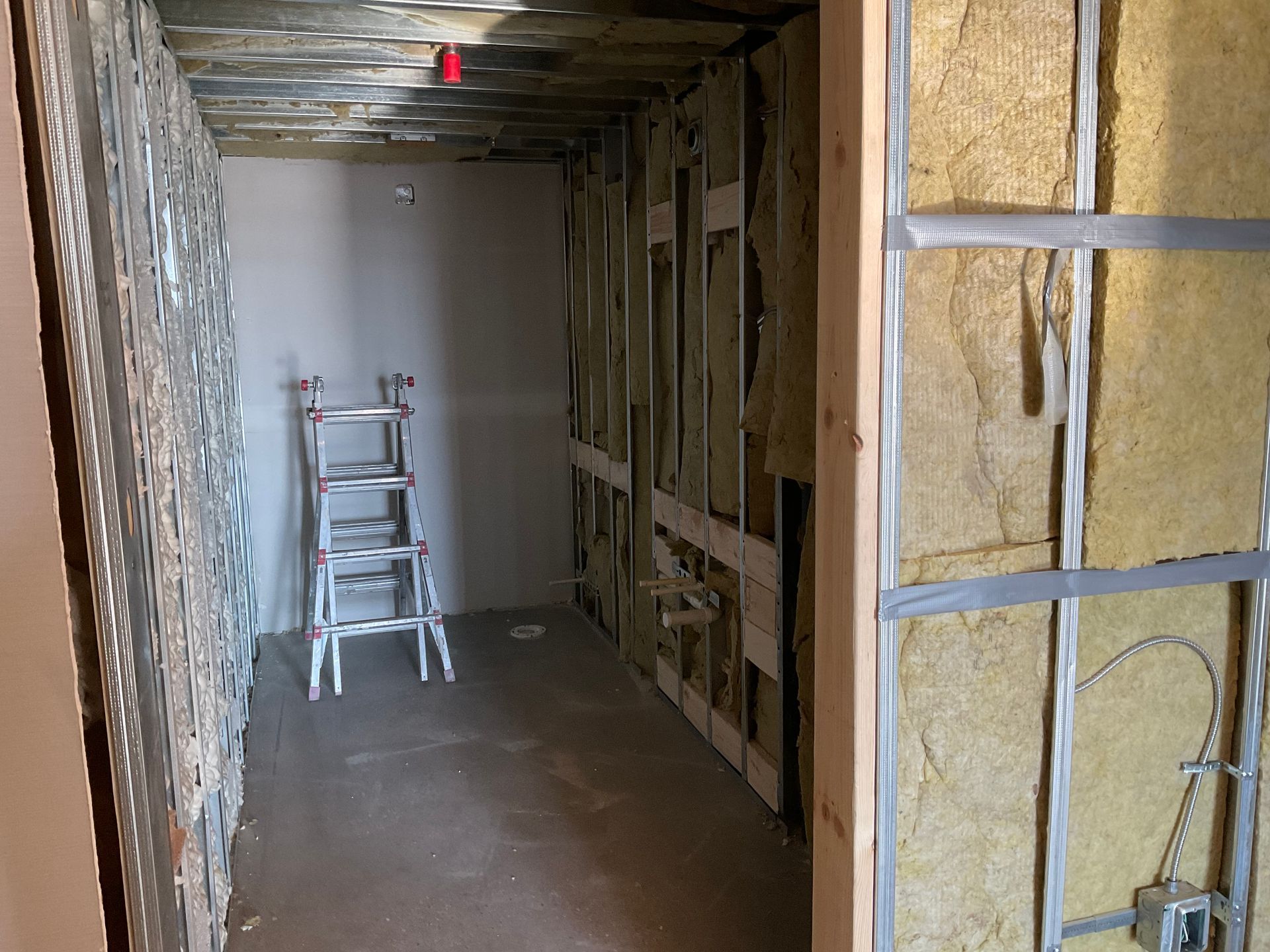 Interior room under construction, metal stud walls, insulation, unfinished ceiling, small ladder.