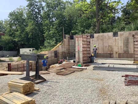 Construction site: workers near concrete walls, gravel ground, trees in background.