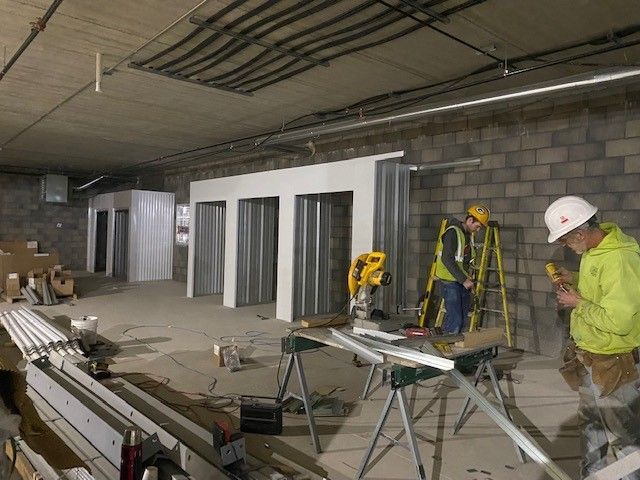 Construction workers installing bathroom stalls in a concrete-walled space, lit with overhead fluorescent lights.