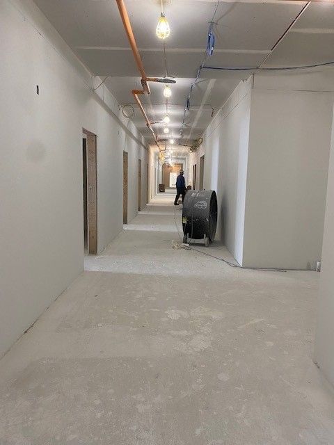 Empty construction hallway with person and large fan. Gray walls and flooring, exposed pipes and lights.