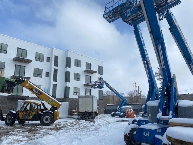 Construction site with snow. White building with balcony, blue lift equipment, yellow forklift, and cloudy sky.