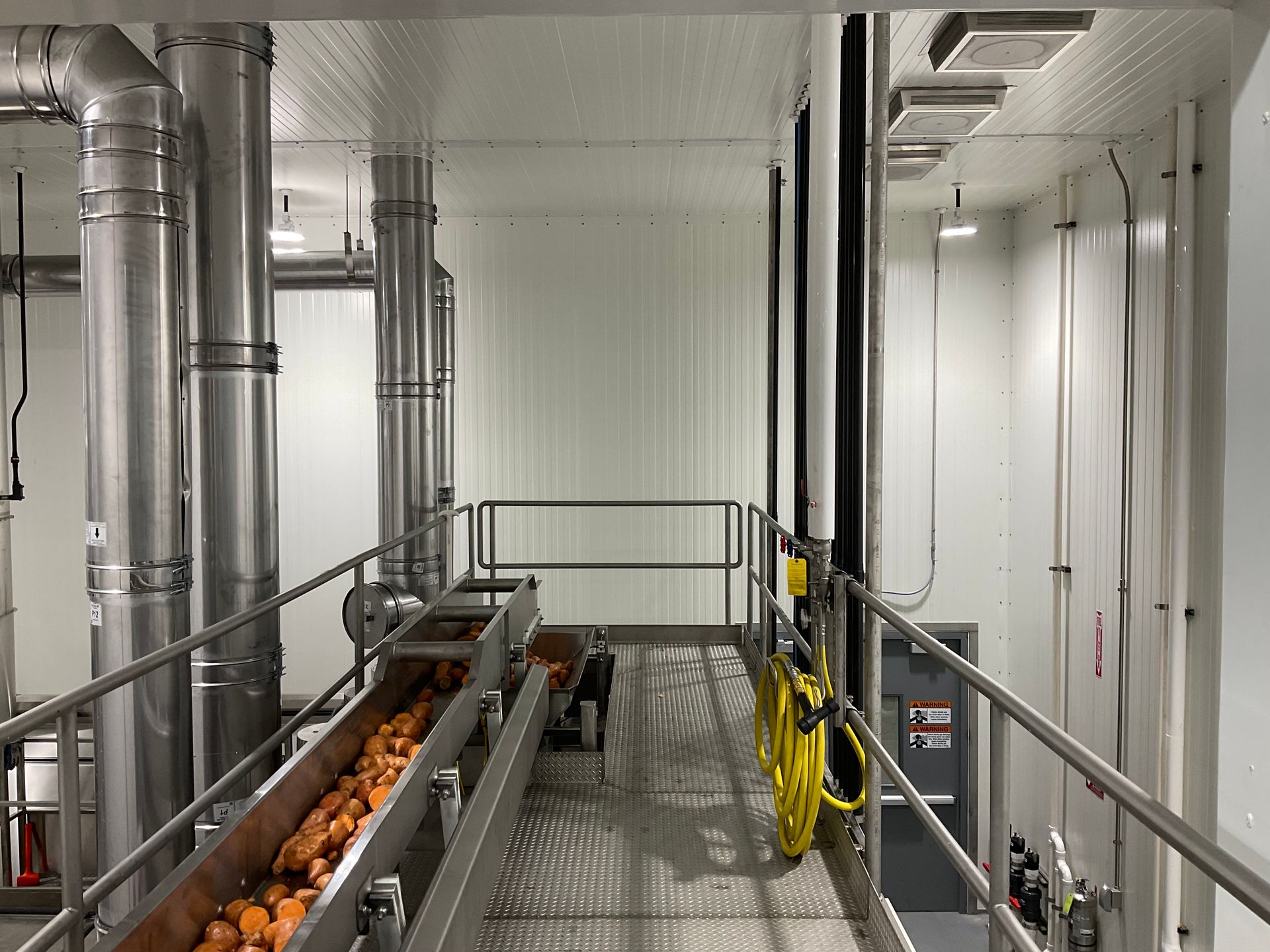 Conveyor belt with produce in a white industrial space, metal pipes, and a walkway.