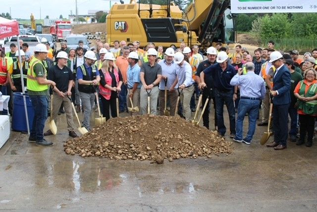 People with shovels around a pile of dirt, likely at a groundbreaking ceremony. A large yellow construction vehicle is in the background.