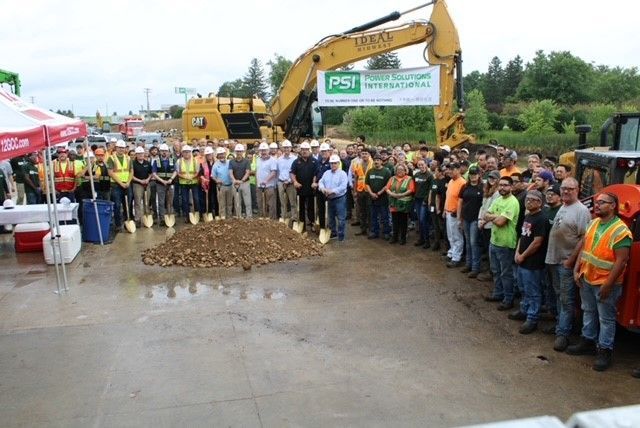 Group of people with shovels at a ground-breaking ceremony next to an excavator.