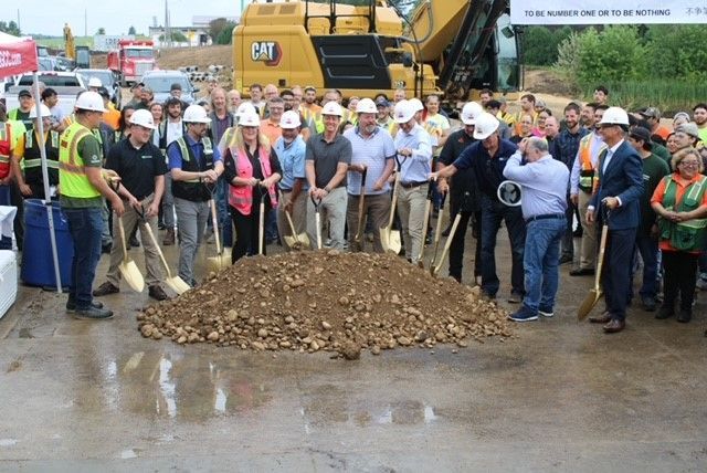People with shovels at a ground breaking ceremony, gathered around a pile of dirt. A large yellow construction vehicle is in the background.