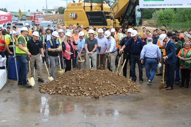 People with shovels around a pile of dirt, likely a groundbreaking ceremony for construction. A large yellow machine is in the background.