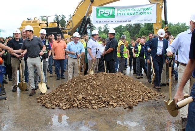 Group of people in hard hats and business attire break ground with shovels near a dirt pile and a construction vehicle.