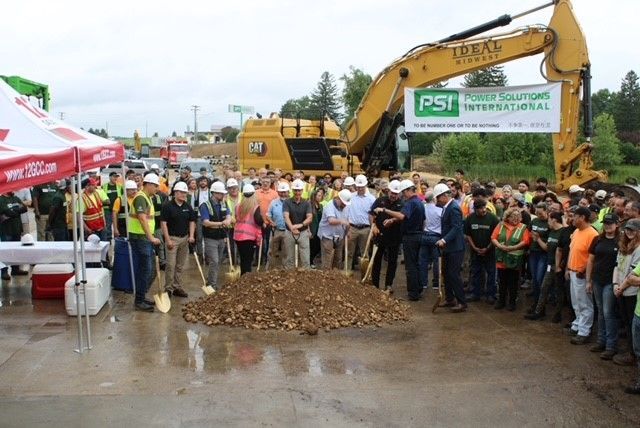 Groundbreaking ceremony with a crowd gathered around a pile of dirt, yellow excavator in background.